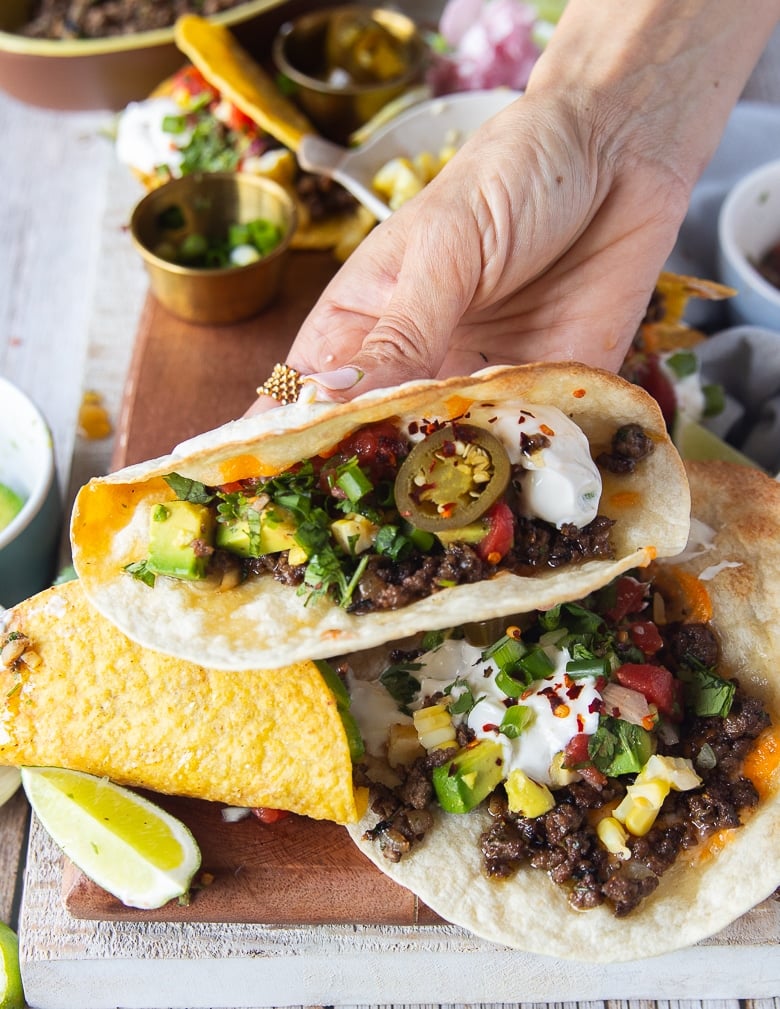 A hand holding a close up of a ground beef tacos showing the inside juicy beef melting cheese and all the topping like sour cream, salsa, cheesy sauce, guacamole and jalapeno